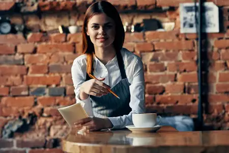 Официантка с чашкой кофе стоит возле стола на фоне кирпичной стены