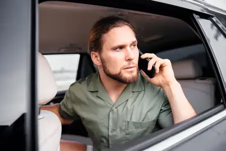 Бородатый длинноволосый мужчина сидит в автомобиле и разговаривает по телефону, демонстрируя сосредоточенность