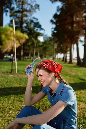 Улыбающаяся женщина в красной бандане и джинсовом наряде сидит на траве в солнечном парке в окружении зелёных деревьев, наслаждаясь тёплым днём с весёлым настроением и естественным светом на улице