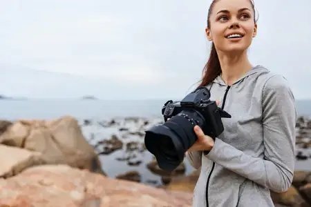 Молодая женщина радостно держит фотоаппарат у океана, одетая в повседневную серую спортивную одежду