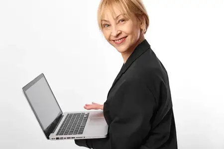 Женщина, ноутбук, бизнес, студийный портрет, улыбающаяся пожилая женщина, черный блейзер, открытый ноутбук, дружелюбное выражение лица, стоит на белом фоне, удаленная работа, онлайн-общение