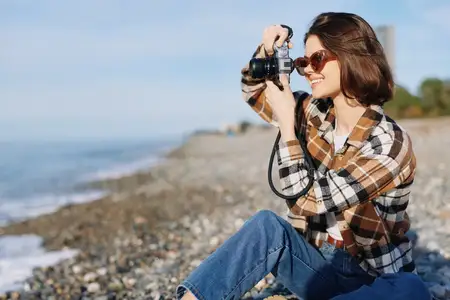 Непринужденная сцена пляжной фотосъемки с улыбающейся женщиной-фотографом, фотоаппаратом в руках, джинсами, клетчатой рубашкой, солнечными бликами, морским пейзажем и творческими образами на…