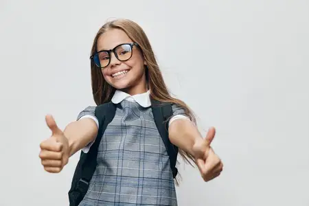 Улыбающаяся школьница в очках и с рюкзаком показывает жест «класс», уверенный счастливый ребёнок в форме изолирован на светлом фоне, символизируя учёбу и достижения