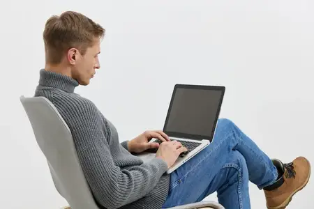 Молодой человек работает за ноутбуком, одетый в серую водолазку и синие джинсы, сидит на современном стуле, печатает на компьютере, сосредоточенный, концепция удаленной работы, образ жизни цифрового…