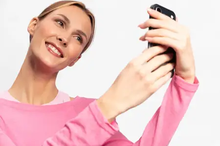 Счастливая женщина в розовом топе держит смартфон и делает селфи на белом фоне