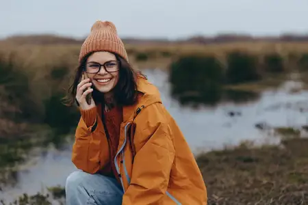Сцена отдыха на природе: улыбающаяся женщина в оранжевой куртке и вязаной шапке разговаривает по телефону на фоне сельского пейзажа