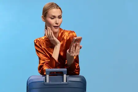 Молодая женщина в оранжевой атласной рубашке держит смартфон с удивленным выражением лица, опираясь на синий дорожный чемодан