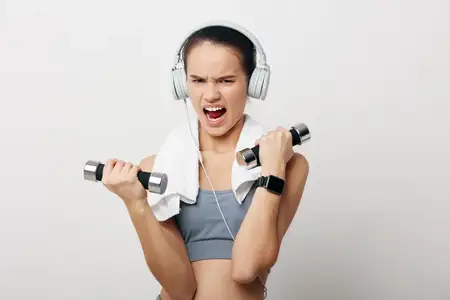 Спортивная женщина с решимостью выполняет сгибания рук с гантелями в наушниках на фоне, напоминающем тренажерный зал