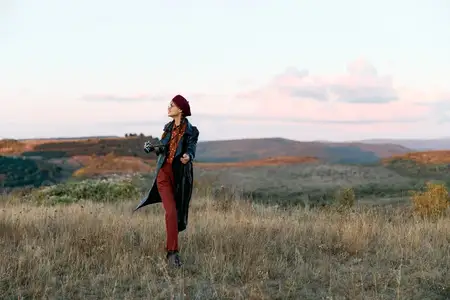 Умиротворенная женщина в эффектном красном пальто грациозно стоит в бескрайнем поле в окружении захватывающих дух гор вдалеке