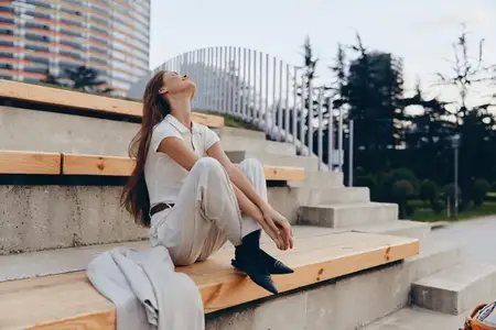 Деловая женщина наслаждается моментом размышления на уличных ступенях, одетая в белое, излучая уверенность и спокойствие на фоне яркого городского пейзажа