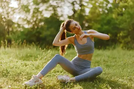 Спортивная женщина в серой спортивной одежде выполняет растяжку в солнечном парке, демонстрируя решимость и уверенность