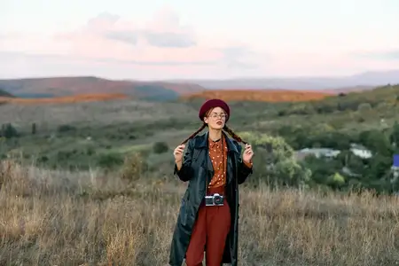 Уверенная женщина в черном пальто и красных брюках принимает позу в бескрайнем поле в сумерках, излучая загадочность и элегантность