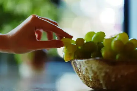 Рука тянется к миске со свежим виноградом на кухонной стойке, на заднем плане видно растение