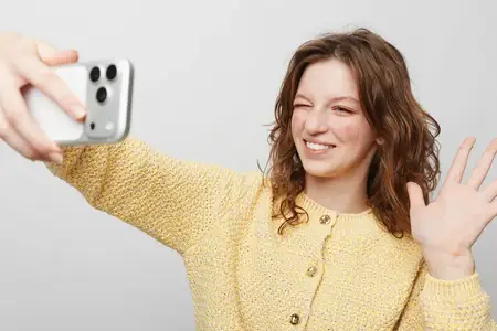 Улыбающаяся девушка в вязаном желтом свитере делает селфи на телефон, махая рукой в яркой студии