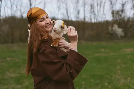 Молодая женщина в коричневом пальто и жёлтом платке нежно держит на руках маленькую козу на травянистом поле с голыми деревьями на заднем плане в облачный день