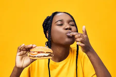 Радостная молодая женщина африканского происхождения наслаждается бургером в яркой жёлтой обстановке, что подчёркивает удовольствие от еды и энергичный образ жизни