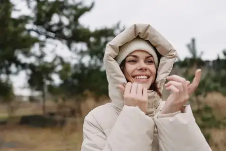 Улыбающаяся женщина в утепленной куртке с капюшоном наслаждается свежим воздухом и природным пейзажем, выражая комфорт и радость в холодное время года