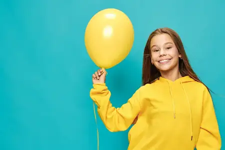 Улыбающаяся девочка в желтой толстовке держит воздушный шарик на фоне голубого фона