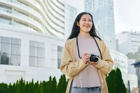 Улыбающаяся женщина с фотоаппаратом в городской среде, на улице, на фоне стеклянных башен