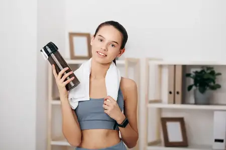 Спортивная женщина в спортивном топе и леггинсах с бутылкой воды, стоящая в минималистичном интерьере с декором и полками, передающая мотивацию, тренировки и здоровье во время занятий