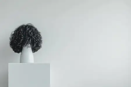 Этот художественный манекен с кудрявыми волосами демонстрирует современную прическу на минималистичном пьедестале на мягком сером фоне, идеально подходит для концепций моды или красоты