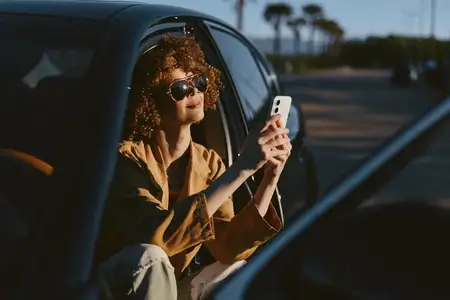 Женщина с вьющимися волосами в машине, держит смартфон, в солнцезащитных очках, цвет ретро-пленки, повседневный модный образ, непринужденная сцена образа жизни, естественный свет и теплое сияние