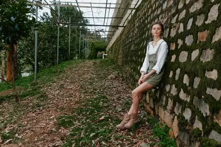 Молодая женщина европейской внешности в светлой блузке и комбинезоне сидит у каменной стены, увитой плющом, в окружении пышной зелени, вызывая чувство спокойствия и умиротворения