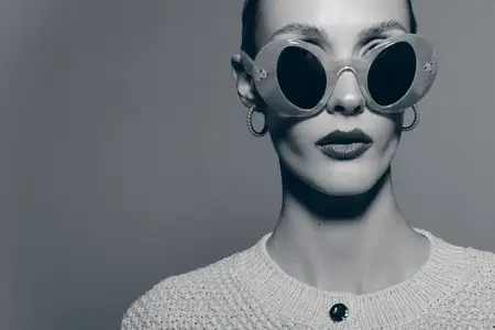 Крупный план светлокожей женщины в круглых солнцезащитных очках и серьгах-кольцах, одетой в фактурный свитер на однотонном монохромном фоне