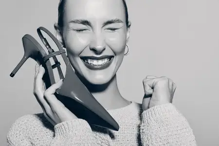 Улыбающаяся белая женщина со светлой кожей с туфлями на высоком каблуке возле лица, выражающая счастье и модный стиль в студийной обстановке