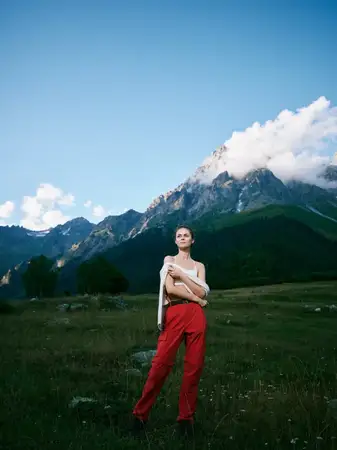 Альпийская сцена с горами, лугом и ясным небом; женщина в красных брюках позирует среди полевых цветов, запечатлевая свободу, исследования и красоту жизни на открытом воздухе