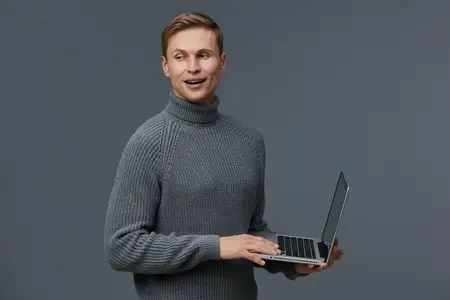 Радостный мужчина с короткими каштановыми волосами, одетый в теплый серый свитер с горлом, стоит на нейтральном сером фоне, держа ноутбук, и выражает счастье, удовлетворение или успех
