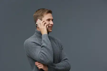 Счастливый молодой человек с короткими светлыми волосами, одетый в уютный серый свитер с высоким воротником, держит смартфон у уха и широко улыбается