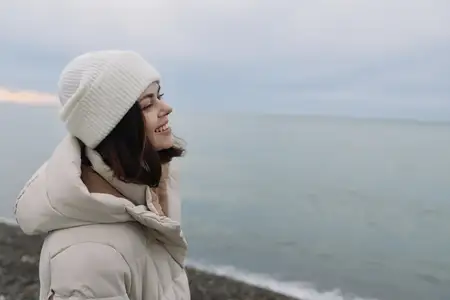 Женщина в теплом зимнем пальто и вязаной шапке смотрит в сторону моря