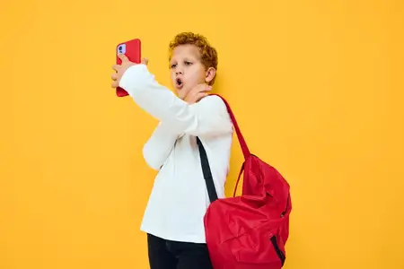 Забавный мальчик-подросток с красным рюкзаком разговаривает по телефону на жёлтом фоне