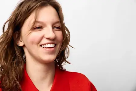 Счастливая молодая женщина с вьющимися каштановыми волосами в красной одежде мягко улыбается на белом фоне