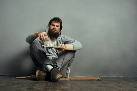 Бездомный мужчина сидит на полу, держит табличку с просьбой о помощи и курит сигарету