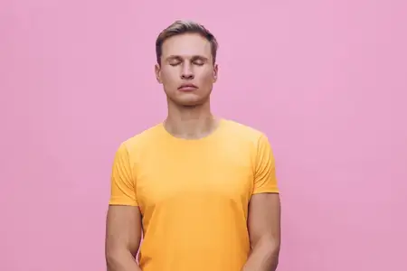 Спокойный молодой человек с короткими волосами, одетый в желтую футболку, медитирует с закрытыми глазами в студийной обстановке на розовом фоне, минималистичная композиция, безмятежная и мирная…