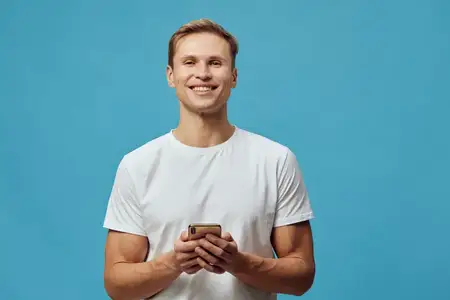 Счастливый улыбающийся молодой человек европейской внешности в белой футболке держит смартфон и смотрит в камеру, изолированно на синем фоне, с положительными эмоциями