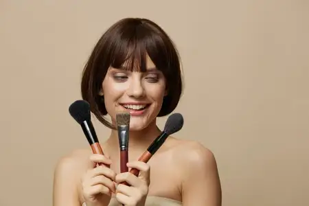 Брюнетка с кисточкой для косметики, привлекательным взглядом позирует на бежевом фоне