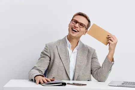 Веселый светлокожий мужчина работает цифровым кочевником, держит конверт, улыбается, носит очки и повседневный пиджак за офисным столом с ноутбуком и телефоном на столе, изолированный на белом фоне