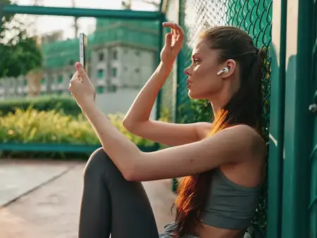 Молодая женщина сидит у зеленого забора, пользуясь смартфоном с беспроводными наушниками, в окружении природы в современной городской среде