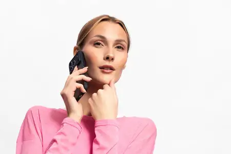 Задумчивая молодая женщина в розовом топе пользуется смартфоном, подперев подбородок рукой