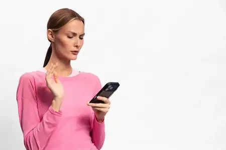 Женщина в розовом свитере держит смартфон и поднимает руку с обеспокоенным выражением лица на белом фоне