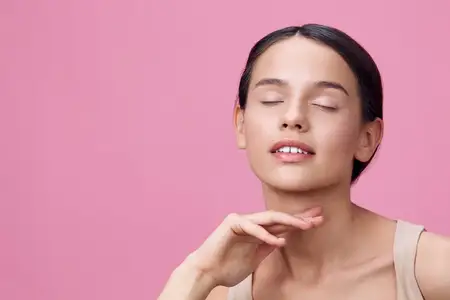 Безмятежная молодая женщина с закрытыми глазами и нежной улыбкой на фоне мягкого розового фона, воплощающая естественную красоту, безмятежность и минималистичную эстетику
