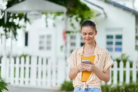 Весёлая молодая женщина увлечённо смотрит в смартфон, стоя на улице рядом с ярким белым домом и штакетным забором