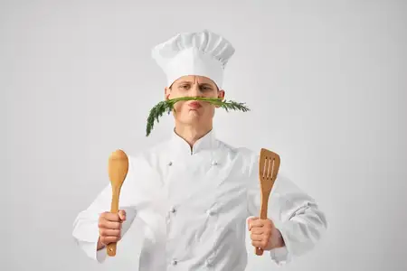 Повар держит кухонную утварь рядом с лицом, на фоне зелени, в ресторане