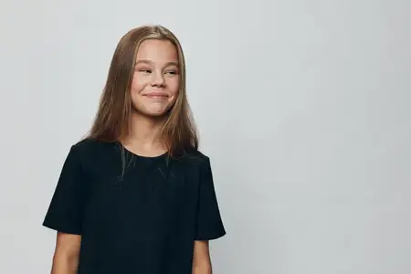 Юная девушка в черной футболке стоит на светлом фоне, смотрит в сторону и улыбается, передавая расслабленное настроение в студийной обстановке