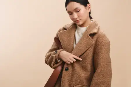 Азиатская женщина в уютном пальто oversize поправляет лямку сумки, поза грациозна, нейтральная обстановка намекает на шикарный профессионализм и расслабленную городскую моду