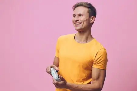 Улыбающийся молодой мужчина в желтой футболке держит игровой контроллер и смотрит в сторону, изолированный на однотонном розовом фоне