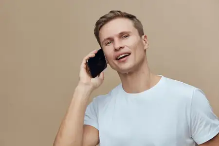 Счастливый мужчина со светло-каштановыми волосами в белой футболке улыбается, прижимая мобильный телефон к уху, и стоит на фоне нейтральной бежевой стены